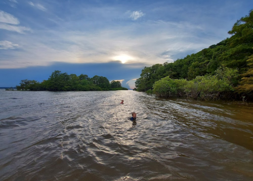Two people swim in a calm river surrounded by lush greenery under a dramatic sky at sunset.