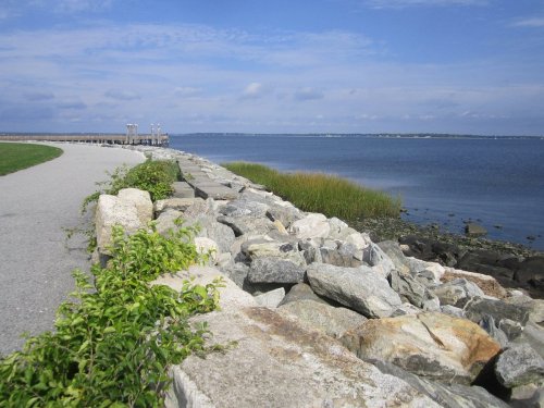 A rocky shoreline with a grassy path, leading to a calm blue sea under a partly cloudy sky.