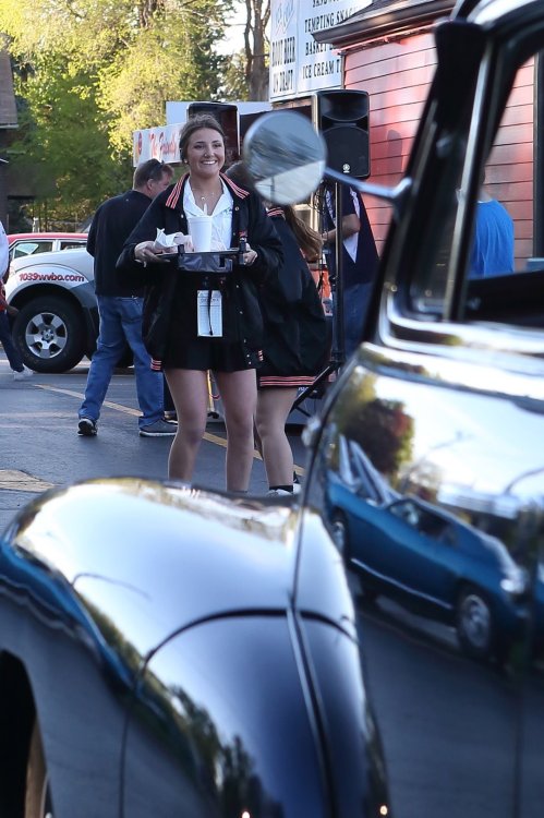 A smiling young woman in a black and red outfit holds a tray, with cars and people in the background.