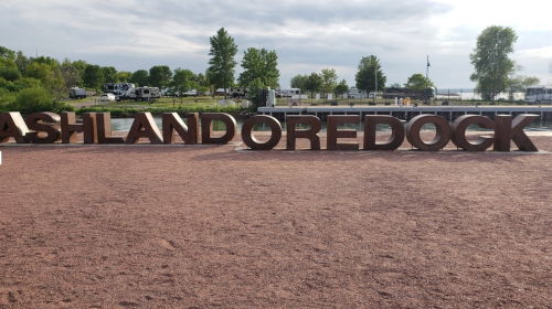Large letters spelling "ASHLAND OREDOCK" on a gravel surface near a waterfront with trees in the background.