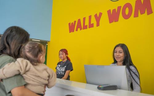 A reception area with a bright yellow wall, two women at the counter, and a person holding a baby.