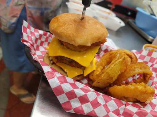 A towering burger with cheese and fried chicken, served with crispy onion rings on a checkered paper plate.