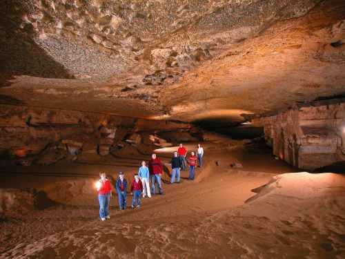 A group of people exploring a large cave, illuminated by flashlights, with rocky walls and a high ceiling.