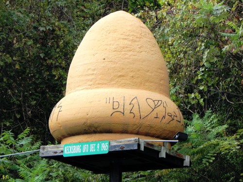 A large, conical structure with graffiti, mounted on a signpost in a wooded area.