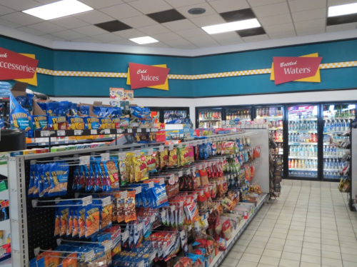 A convenience store aisle filled with snacks, drinks, and a cooler displaying bottled waters. Brightly colored signage above.