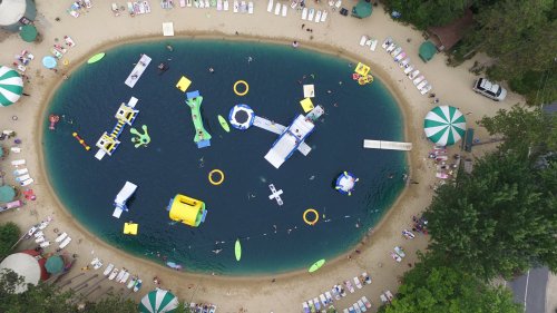Aerial view of a sandy beach with a swimming area, featuring inflatable water toys and sunbathers on lounge chairs.