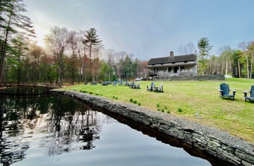 A serene lakeside view featuring a house, green lawn, and Adirondack chairs surrounded by trees.