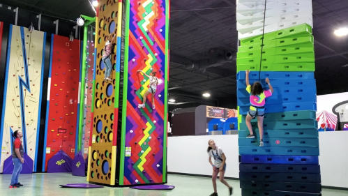 Two colorful climbing walls in an indoor gym, with people climbing and others observing. Bright, playful atmosphere.