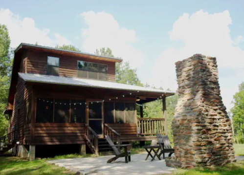 A rustic wooden cabin with a stone chimney, surrounded by trees and a clear blue sky.