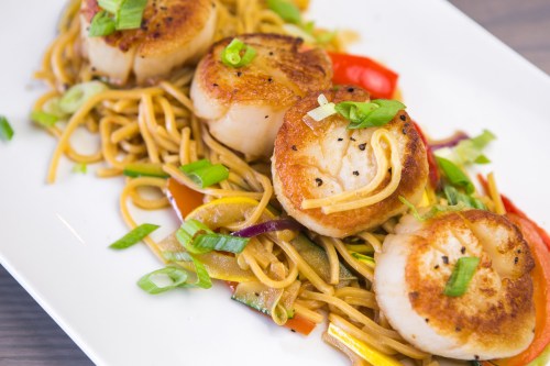Three seared scallops on a bed of stir-fried noodles and colorful vegetables, garnished with green onions.
