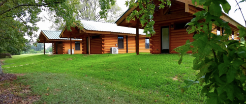 Three log cabins with green lawns and trees, set in a peaceful outdoor environment.