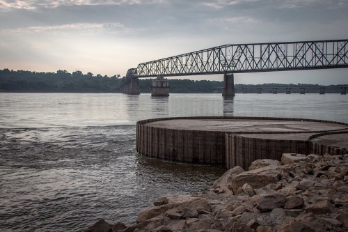 A river scene featuring a bridge in the background, rocky shoreline, and calm water under a cloudy sky.