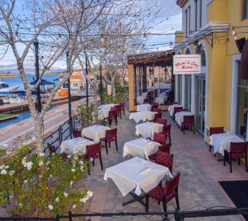 Outdoor dining area with empty tables, white tablecloths, and a view of the water and boats in the background.