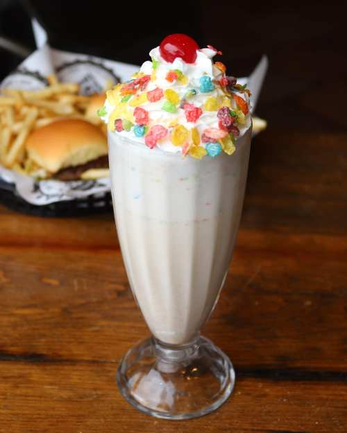 A tall milkshake topped with whipped cream, colorful cereal, and a cherry, with a burger and fries in the background.