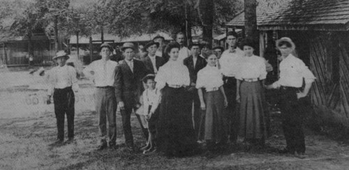 A black-and-white photo of a group of people in early 1900s attire, standing outdoors near trees and buildings.