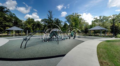 A playground with unique climbing structures and two pavilions, surrounded by trees and blue skies.