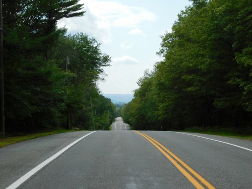 A long, straight road lined with trees under a cloudy sky, stretching into the distance.