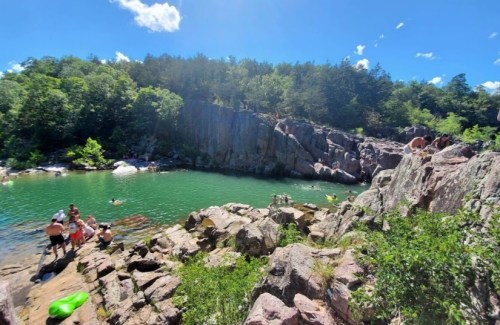 A sunny day at a rocky swimming area with people enjoying the water and lush greenery surrounding the scene.