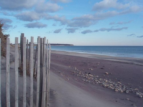 A serene beach scene with wooden fencing, soft sand, and calm blue waters under a partly cloudy sky.