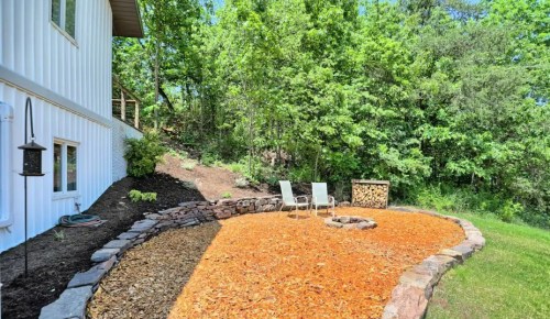 A landscaped outdoor area with two chairs, a fire pit, and a circular bed of wood chips surrounded by greenery.