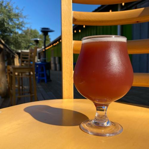 A glass of amber beer sits on a table, with a sunny outdoor setting and wooden chairs in the background.
