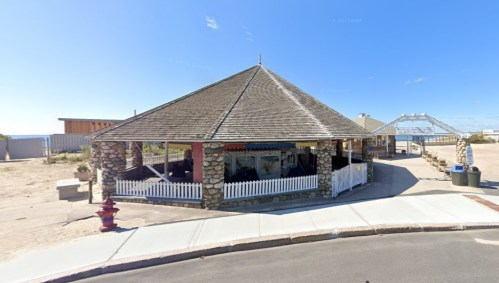 A stone pavilion with a thatched roof, surrounded by a white picket fence, set against a clear blue sky.