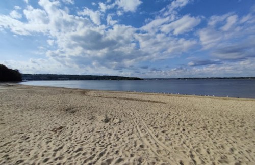 A sandy beach with calm water and a cloudy blue sky in the background.