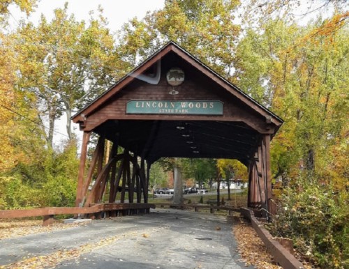 A wooden covered bridge entrance to Lincoln Woods State Park, surrounded by colorful autumn foliage.