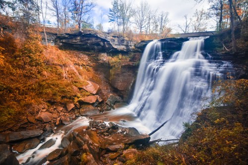 A serene waterfall cascades over rocky cliffs, surrounded by autumn foliage and a cloudy sky.