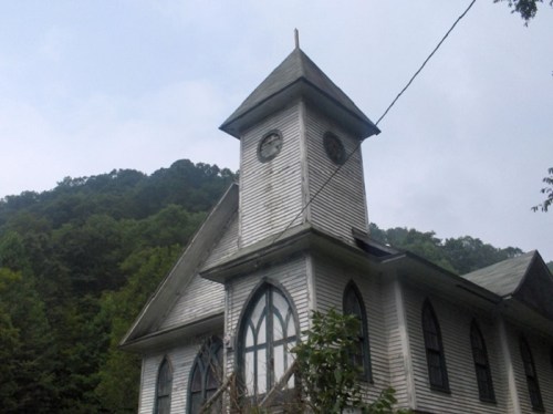 An old church with a tall steeple, surrounded by trees and mountains under a cloudy sky.