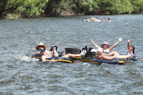 A group of people floating on inner tubes in a river, enjoying a sunny day and waving at the camera.