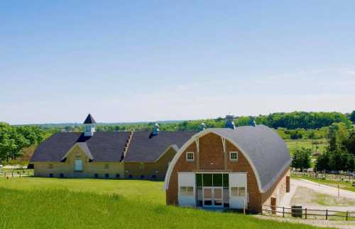 A scenic view of two large barns surrounded by green fields and trees under a clear blue sky.