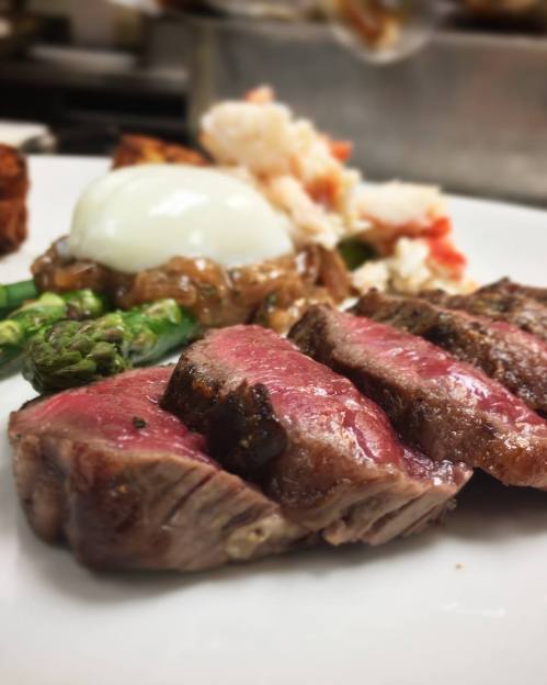 A plate featuring sliced steak, asparagus, a poached egg, and a seafood dish, beautifully arranged.