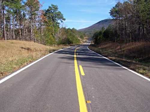 A winding road stretches through a forested landscape, with hills in the background under a clear blue sky.