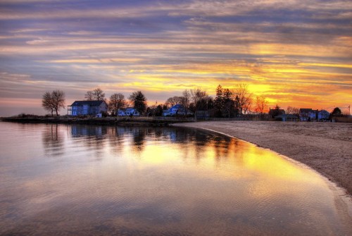 Sunset over a calm beach, reflecting colorful skies and silhouettes of trees and houses along the shoreline.