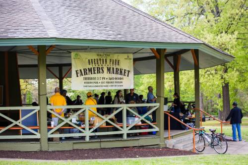 A farmers market under a gazebo with a banner, people browsing stalls, and a bicycle parked nearby.
