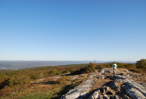 A person walks along a rocky outcrop, overlooking a vast landscape of rolling hills and clear blue sky.