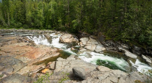 A serene river flows over rocky terrain, surrounded by lush green trees and a peaceful natural landscape.