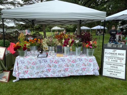 A flower stand at an outdoor market, featuring colorful blooms in buckets on a floral tablecloth under a white tent.