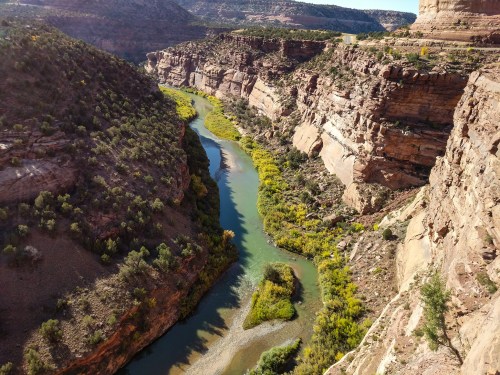 A winding river flows through a rocky canyon, surrounded by lush greenery and steep cliffs under a clear blue sky.