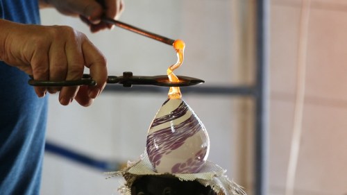 A person shapes molten glass into a decorative piece using tools, with vibrant colors swirling in the glass.