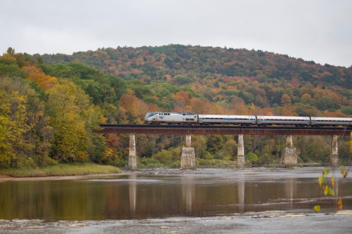 A train crosses a bridge over a river, surrounded by vibrant autumn foliage and rolling hills.