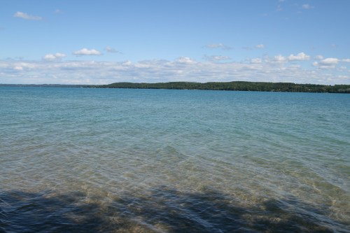 A serene lake with clear blue water and a distant green shoreline under a partly cloudy sky.