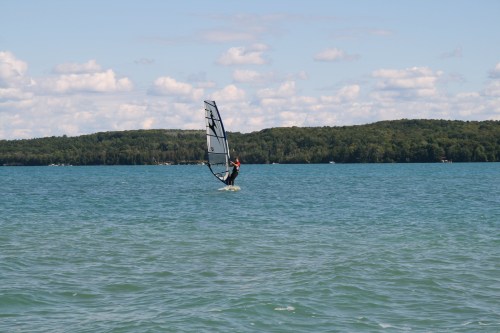 A windsurfer glides across a calm lake with green hills in the background under a partly cloudy sky.