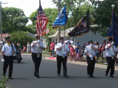 A parade scene with flag bearers in uniform, marching down a street lined with spectators.