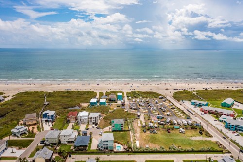 Aerial view of a beach with colorful houses, parked cars, and a vast ocean under a partly cloudy sky.