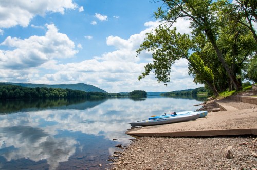 A calm river scene with a kayak on the shore, surrounded by trees and mountains under a blue sky with fluffy clouds.