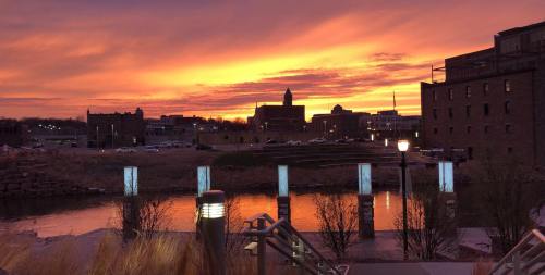Sunset over a city skyline, reflecting on water, with silhouettes of buildings and glowing streetlights.