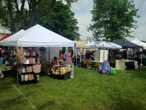 Two market stalls with white and black tents, displaying various handmade goods in a grassy area.