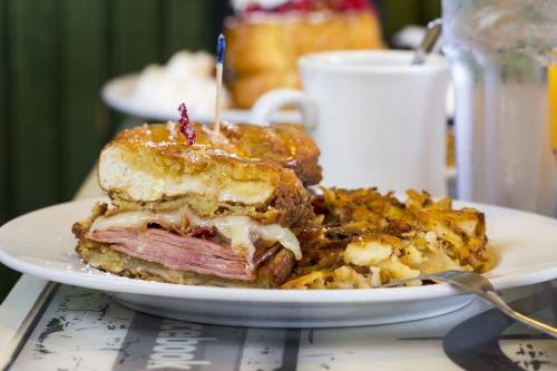 A plate featuring a sandwich made with French toast, ham, and cheese, alongside crispy hash browns and a coffee cup.